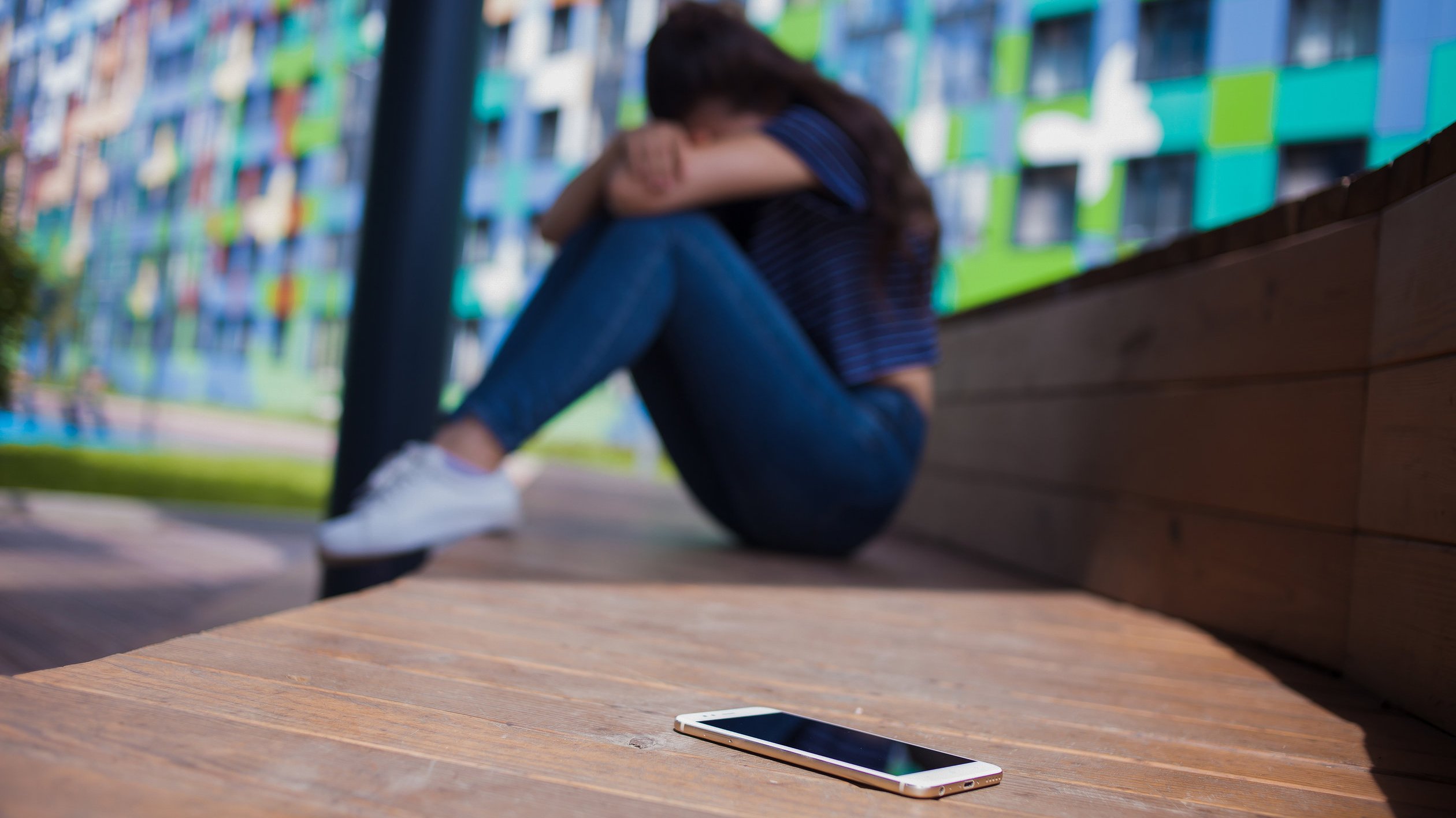 Smartphone lying in the foreground, on blurred background, girl, hiding face in her knees, sitting on a wooden bench.